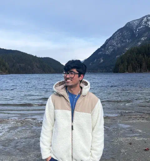 Apram in a white fleece jacket smiling on a lakeshore, with forested, snow-dusted mountains behind him