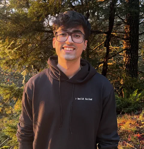 Apram in a dark hoodie and glasses smiling in a forest lit by low evening sun